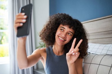 Doğal kıvırcık saçlı neşeli bir kadın akıllı bir telefonla selfie çekiyor. Parlak bir şekilde gülümsüyor ve barış işareti gösteriyor. Fotoğraf, konforlu bir ev ortamında pozitifliği ve modern bir yaşam tarzını yakalıyor..