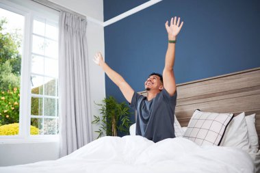 Neşeli genç bir adam aydınlık ve havadar bir yatak odasında uyanır, rahat bir şekilde güne başlar. Sabah güneşi misafirperver atmosferi güçlendirir..