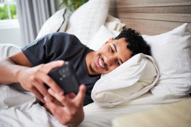 Neşeli bir yüzü olan, yatakta rahat bir şekilde uzanan bir adam, akıllı telefonunu kullanır. Aydınlık ve rahat ortam rahatlamayı ve evde hoş bir sabah anını ön plana çıkarıyor..