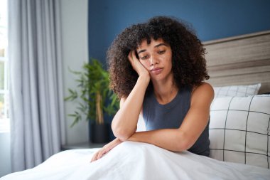 Kıvırcık doğal saçlı genç bir kadın huzurlu ve aydınlık bir yatak odasında bir yatağa oturur. Kafası elinde dinlenirken yansıtıcı görünüyor. Parlak doğal ışık odayı aydınlatır..