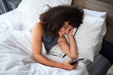 Doğal kıvırcık saçlı genç bir kadın rahat bir yatakta uzanıyor, akıllı telefonunu neşeli bir ifadeyle kullanıyor. Dekor yumuşak bir şekilde aydınlatılmış, rahat ve rahat bir atmosfer sergiliyor..