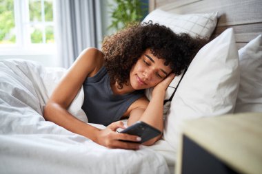 Etrafında yastıklar ve akıllı telefon kullanan bir gencin rahat bir şekilde yatakta yattığı sakin bir sabah sahnesi. Oda doğal gün ışığıyla aydınlanmış, sıcak atmosfere katkı sağlıyor..