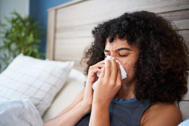 Genç bir kadın, semptomlarını gidermek için mendil kullanarak modern bir evde yatağında oturuyor. İfadesi rahatsızlık veriyor, hastalık ve kendine bakım konularını vurguluyor..