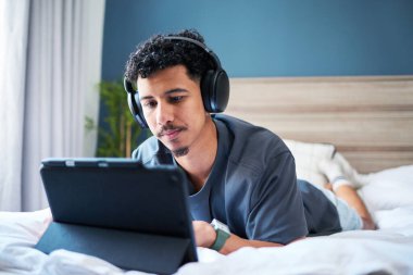 Sıcak bir odada yatakta uzanmış, kulaklık takmış, tabletle meşgul olan bir adam. Huzurlu ortam, rahat bir ev ortamında kişisel rahatlama ve modern yaşam tarzı faaliyetlerini vurguluyor..