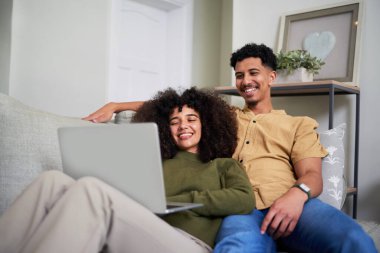 Mutlu bir çift rahat bir kanepede rahatlar, rahat evlerinde dizüstü bilgisayarda gezinirken kahkahalarını paylaşırlar. Ortam sıcak ve davetkar, birlikte geçirdiğimiz kaliteli zamanı yansıtıyor..