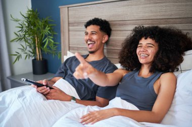 Neşeli bir çift yatakta oturup bir televizyon programının keyfini çıkarıyor. Neşeli ifadeleri rahatlamayı ve samimi, modern bir ev ortamında kahkahaları paylaşmayı yansıtıyor, arkadaşlığı ve eğlenceyi vurguluyor..