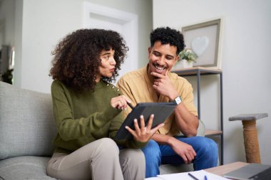 Kanepede oturan genç bir çift dijital tablete bakarken neşeli bir anı paylaşıyorlar. Sıcak, davetkar alan arkadaşlığı ve günlük aktiviteleri yansıtıyor, neşeli bir atmosferi teşvik ediyor..