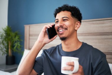 Neşeli bir adam hoş bir telefon konuşması yaparken elinde bir fincan kahve tutarken görülüyor. Minimalist dekor ve parlak, modern bir atmosferi olan rahat bir yatak odasında oturuyor, pozitifliği yansıtıyor..