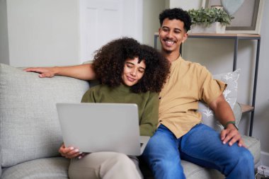 Neşeli bir çift, evlerindeki koltuklarında oturmuş birlikte vakit geçiriyorlar. Rahatlamış görünüyorlar ve bir dizüstü bilgisayara bakıyorlar, rahat ve iyi dekore edilmiş bir yerde keyifli bir anı paylaşıyorlar..