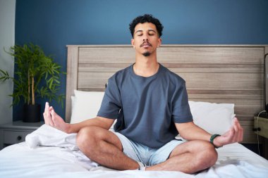 Yatak odası ortamında meditasyon yapan genç bir adam sükunet ve kendine bakmanın tadını çıkarıyor. Huzurlu ortam farkındalık, sağlık ve dengeli bir yaşam tarzına odaklanmayı tamamlar..