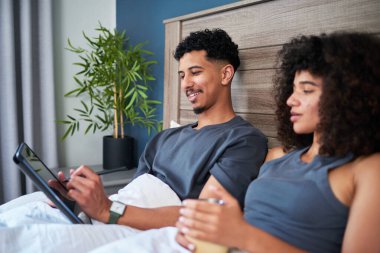 Genç bir çift yatakta uzanıyor, sessiz anlarını paylaşıyor ve dokunmatik ekran cihazları ve içecekleriyle meşgul oluyor, rahatlamayı, dijital bağlantıyı ve huzurlu bir ev ortamını sembolize ediyor..