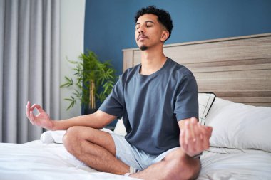 Evde farkındalık pratiği yapan bir adam, sakin bir yatak odası ortamında meditasyon yapıyor. Yatakta bacak bacak üstüne atarak rahatlamaya ve zihinsel berraklığa odaklanarak doğal bir dekorasyonla sükuneti arttırıyor..