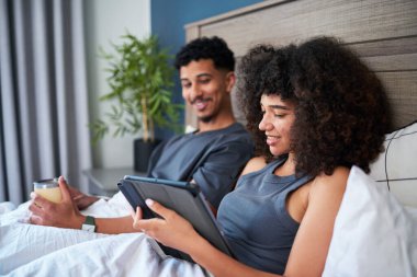 Neşeli bir çift, sakin bir sabahı yatakta geçirir, tablet üzerinde dijital içerikle meşgul olarak, kahve yudumlarken. Sıcak atmosfer rahat bir ev ortamında rahatlığı ve birlikteliği yansıtıyor..