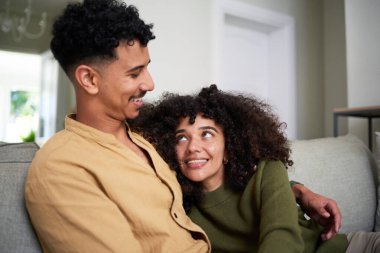 Neşeli bir anı paylaşan genç bir çiftin parlak, rahat bir yuva ortamında koltukta rahat bir şekilde oturduğu pozitiftir. Yerel bir ortamda bağlantı, mutluluk ve rahat birlikteliği vurgular.