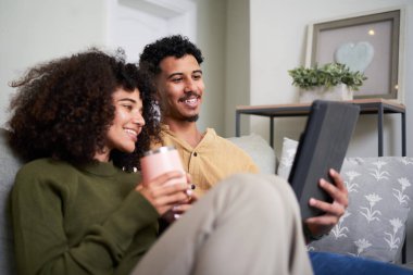 Neşeli genç bir çift, rahat bir oturma odasında birlikte tablet okurken koltuklarında dinlenmekten zevk alır. İfadeleri neşeyi, rahatlığı ve birlikteliği yansıtıyor..