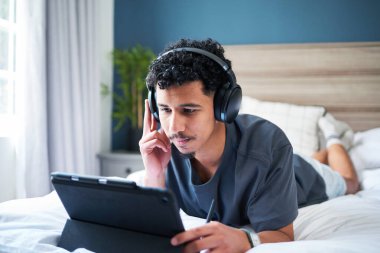 Genç bir adam rahat bir çevreye sahip bir yatakta uzanıyor, kulaklık takıyor ve bir tabletle meşgul. Çevre, o dinlerken ya da cihazındaki içeriği izlerken rahatlama ve odaklanma yayar..