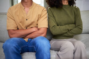 Açık renkli bir kanepede oturan iki kişi, kolları çapraz, gözle görülür mesafede, bir ilişkideki gerginlik veya anlaşmazlığı resmediyor. Görüntü, çatışma ve tefekkür duygularını yakalar..