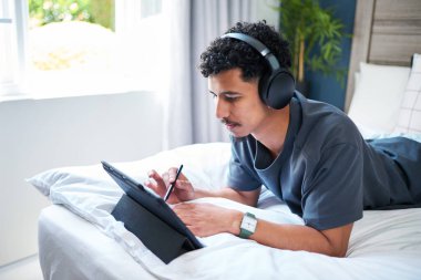 Sanat yaratmak için kulaklık takan genç bir adam, dijital tablet ve stil kullanarak rahat bir şekilde yatağında yatıyor. Ayarlar verimlilik için rahat ve rahat..