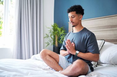 Huzurlu bir yatak odası ortamında bacak bacak üstüne atarak meditasyon yapan bir adam. Odada, gevşemeyi ve iç huzuru artıran yumuşak doğal ışık, rahat dekor ve sakinleştirici unsurlar yer alıyor..