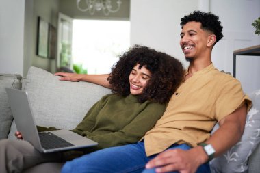 Neşeli bir çift, dizüstü bilgisayar kullanırken keyifli bir anı paylaşarak kanepede dinlenir. Sıcak ve samimi ortam, onların rahat yaşam alanlarında sevgi, bağlantı ve paylaşılan mutluluğu vurgular..