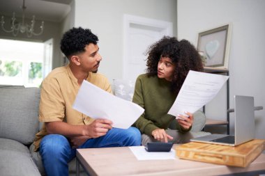 Oturma odalarında oturan genç bir çift ellerinde belgelerle mali konuları tartışıyor. Masada, ev ekonomisi ve önemli kararların yönetiminde takım çalışmasını vurgulayan bir dizüstü bilgisayar bulunuyor..