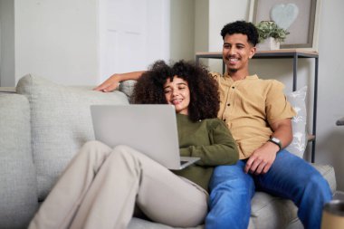 Evdeki kanepede oturmuş laptopa bakarken birlikte vakit geçirmekten zevk alan gülümseyen bir çift. Sıcak ve neşeli bir atmosfer rahatlamayı, mutluluğu ve modern bağlantıyı simgeler..