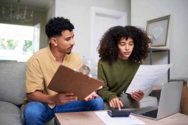 Oturma odasındaki masada finansal planlama üzerine çalışan bir çift. Bir dizüstü bilgisayar ve hesap makinesi kullanarak belgeleri incelerken, bütçe ve hedefleri hakkında tartışıyorlar..