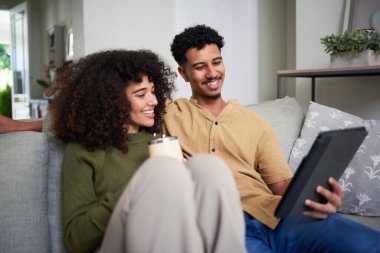 Mutlu genç çift modern evlerindeki kanepede birlikte vakit geçiriyorlar. Gülümsüyorlar, bir tablet tutuyorlar ve sıcak bir içecek içiyorlar, rahat ve rahat bir atmosfer yaratıyorlar..