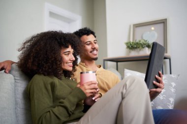 Neşeli bir çift kanepede birlikte oturur, kahve içerken tabletle meşgul olurlar. Konforlu ve misafirperver oturma odası manzaraya sıcaklık katıyor, konfor ve modern bağlılık tasvir ediliyor..