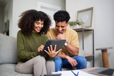 Neşeli bir çift, oturma odalarındaki rahat bir kanepede oturup gülümseme ve kahkahaları paylaşırken tabletle meşgul olurlar. Ortam rahat ve modern, rahat bir atmosferi yansıtıyor..