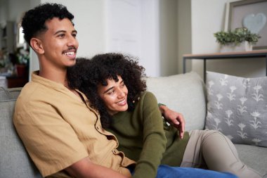 Rahat bir kanepede yan yana oturan genç bir çift şık oturma odalarında sakin ve neşeli bir anın tadını çıkarıyor. Mekan rahatlık, mutluluk ve rahat bir ortam yayar..