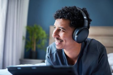 Neşeli genç bir adam rahat bir ev ortamında kablosuz kulaklık kullanarak müzik dinlemekten hoşlanır. Rahatlayan ve meşgul görünen çevresi huzurlu ve rahat bir atmosfer izlenimi veriyor..