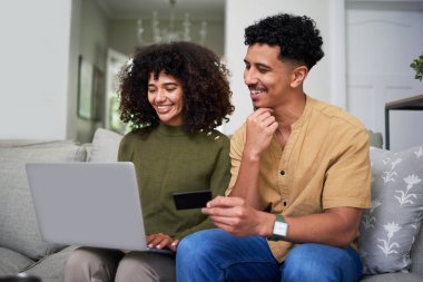Evdeki rahat bir kanepede oturan neşeli bir çift çevrimiçi aktivitelere girişiyor. Aktif olarak bir dizüstü bilgisayar kullanıyorlar ve çevrimiçi alışveriş veya dijital mali işlemlerin altını çizen bir kredi kartı görünür durumda.