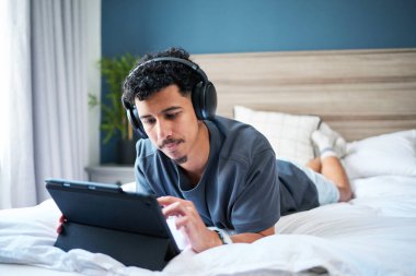 Genç adam yatakta uzanıyor, kulaklık takarken tablet cihazına odaklanmış. Yatak odasının içi sakin ve içine kapanık ifadesi rahatlatıcı ve rahat bir atmosfer sağlıyor..
