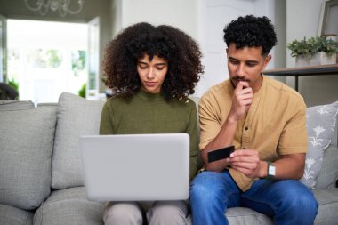 Kanepede oturan genç bir çift, bir dizüstü bilgisayar ve kredi kartı kullanarak düşünceli bir şekilde online alışveriş yapıyor, modern e-ticareti, finansal planlamayı ve günlük hayatta ortak karar alma mekanizmasını simgeliyor..