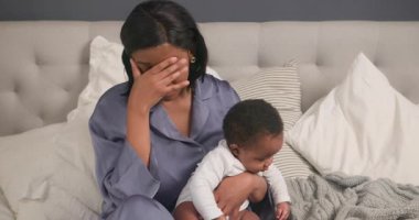 Yorgun bir anne bebeğiyle bir yatakta oturur, şakaklarını ovuşturur, yorgun düşmüştür. Bebek, annesinin yorgunluğu ve stresinden habersiz ayaklarıyla oynar. Yorgun anne, annelik, ebeveynlik ve çocuk bakımı videoları..