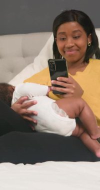 Mutlu bir Afrikalı Amerikalı anne bir yatağa oturur ve telefonuna bakarken bebeğini emzirir. Gülümsüyor ve anın tadını çıkarıyor gibi görünüyor. Bebek rahat görünüyor. Bu sıcaklığı, sevgiyi ve anneliğin neşesini çağrıştırıyor..