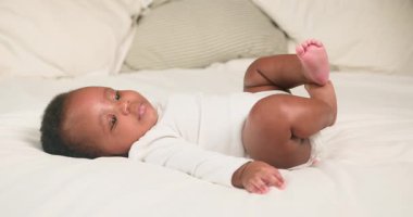 Sevimli Afro-Amerikan bebek bir yatakta sırt üstü uzanıyor, nazikçe bacaklarını tekmeliyor. Bebek mutlu ve rahat görünüyor. Yumuşak ışıklandırma, yumuşak yorgan ve yastıklar rahat ve huzurlu bir atmosfer yaratır..