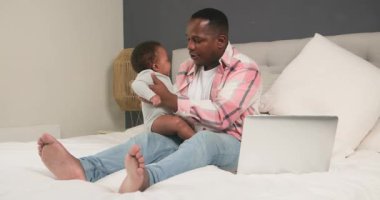 Bir baba dizüstü bilgisayarı ve bebeğiyle yatağında oturuyor. Bebeği kucağında zıplatıyor, onunla etkileşime geçiyor. Baba evde çalışıyor ve bebeğe bakıyor..