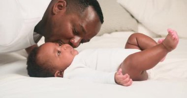 Sevgi dolu bir baba, yatakta yatan bebeğine gülümser. Bebek, babasına bakıyor. Birlikte kaliteli zaman geçiriyorlar. Babası ve çocuğu Afro-Amerikalı. Bu yürek ısıtan sahne, sevgi, bağ ve aileyi çağrıştırıyor..