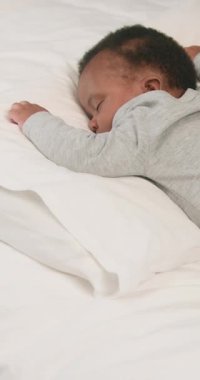 Uyuyan bir bebek, yumuşak, beyaz bir yatakta huzur içinde uzanıp tatlı rüyalar görüyor. Nazik bir el yastığı sevgiyle ayarlar, rahatlık ve dinlenme sağlar. Huzurlu atmosfer, aile içinde huzur, sevgi ve şefkat hissi uyandırıyor..