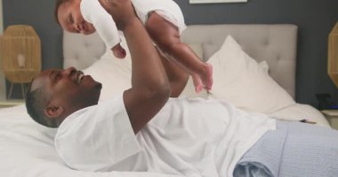 Mutlu bir Afrikalı Amerikalı baba yatağa uzanıyor ve bebeğini havaya kaldırıyor. Bebek beyaz tulum giyiyor ve baba gülümsüyor. Evde ailece çok güzel vakit geçiriyorlar..