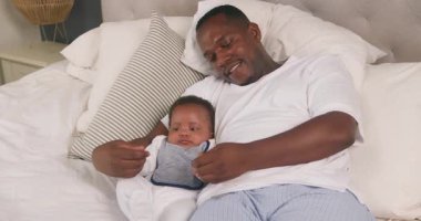 Bir baba ve bebek yatakta yatıyor. Baba beyaz gömlek ve mavi pijama pantolon giyiyor ve bebek de beyaz tulum ve önlük giyiyor. Baba bebeğin ellerini tutuyor ve ona gülümsüyor. Bebek kameraya bakıyor..