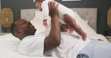 Mutlu bir Afrikalı Amerikalı baba yatakta uzanıyor ve sevimli bebeğini nazikçe havaya kaldırıyor. Babayla çocuk arasındaki sevgi bağı, sıcak evlerinde sevgi dolu bir aile bağı ve neşeli bir neşe yaratıyor..