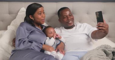 Mutlu bir aile yatakta kaliteli vakit geçirmekten hoşlanır, selfie çeker ve sevimli bebekleriyle anılar yaratır. Ebeveynler, çocuklarıyla etkileşime girerken sevgi ve neşe saçıyor ve telefonlarındaki değerli anları yakalıyorlar..