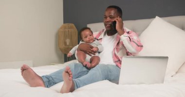 Genç bir baba, evde çalışırken sevimli bebeğiyle iyi vakit geçirmekten hoşlanır. Bir iş görüşmesini ve dizüstü bilgisayarın sorunsuz çalışmasını dengeliyor, rahat bir yatak odası ortamında modern ebeveynlik ve iş hayatı entegrasyonunu gösteriyor..