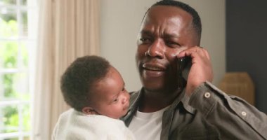 Mutlu bir Afrikalı Amerikalı baba akıllı telefonuyla konuşurken sevimli bebeğini tutarak birden fazla iş yapar. İş ve aile hayatını rahat evinden dengeler, neşe ve bağlantı saçar..