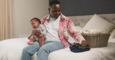 Sevgi dolu bir baba yatak odasında çamaşır katlarken bebeğini kucağına alır. Giysileri sınıflandırıyor ve düzenliyor, çocuk bakımı ve ev hayatının hassas bir anını gösteriyor. Bu iç açıcı sahne ailenin ve sorumluluğun özünü yansıtıyor..