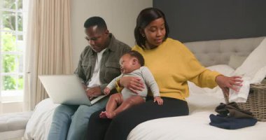 Genç bir aile, yatak odalarında iş ve çocuk bakımını dengeler. Babası dizüstü bilgisayarında çalışıyor, annesi bebek kıyafetlerini katlıyor ve bebekleri annesinin kucağına oturuyor. Modern ebeveynliği, evden çalışmayı ve aile hayatını temsil ediyorlar..