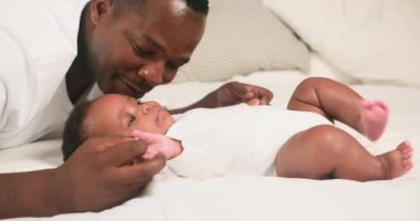 Sevgi dolu bir baba gülümser ve bebeğinin elini nazikçe bir yatakta tutar. Beyaz tulum giyen bebek, şakacı bir şekilde bacaklarını tekmeliyor. Bu iç açıcı sahne ebeveyn ve çocuk arasındaki hassas bağı yakalar..